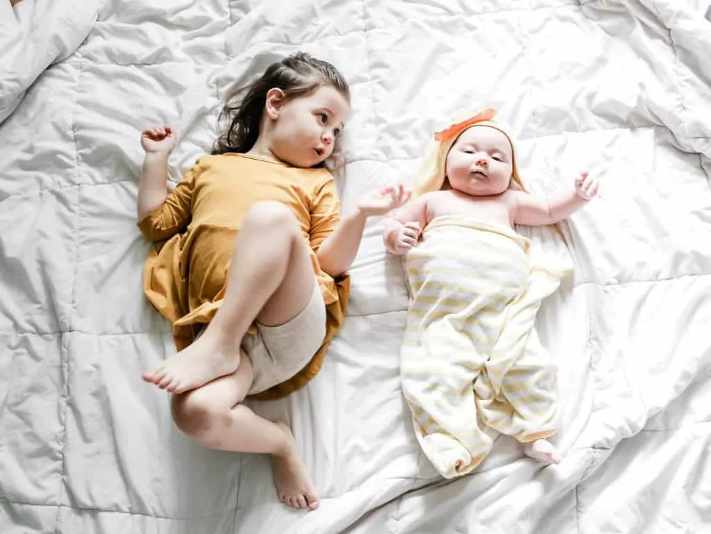 Toddler girl lays next to baby on bed after bath time.