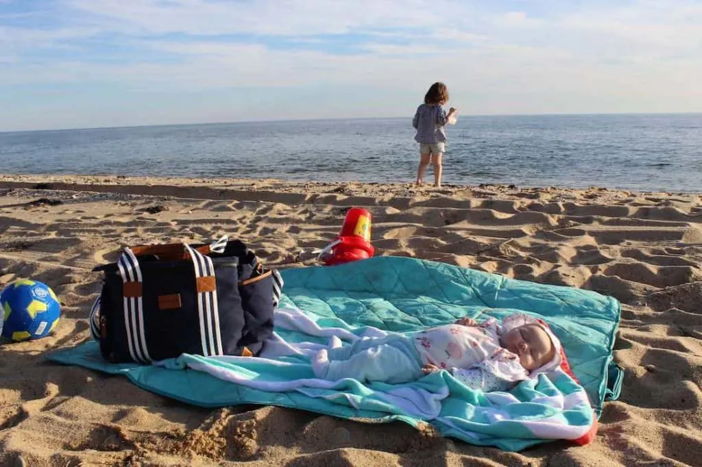 Baby sleeps on blanket on beach with toddler girl playing in background.