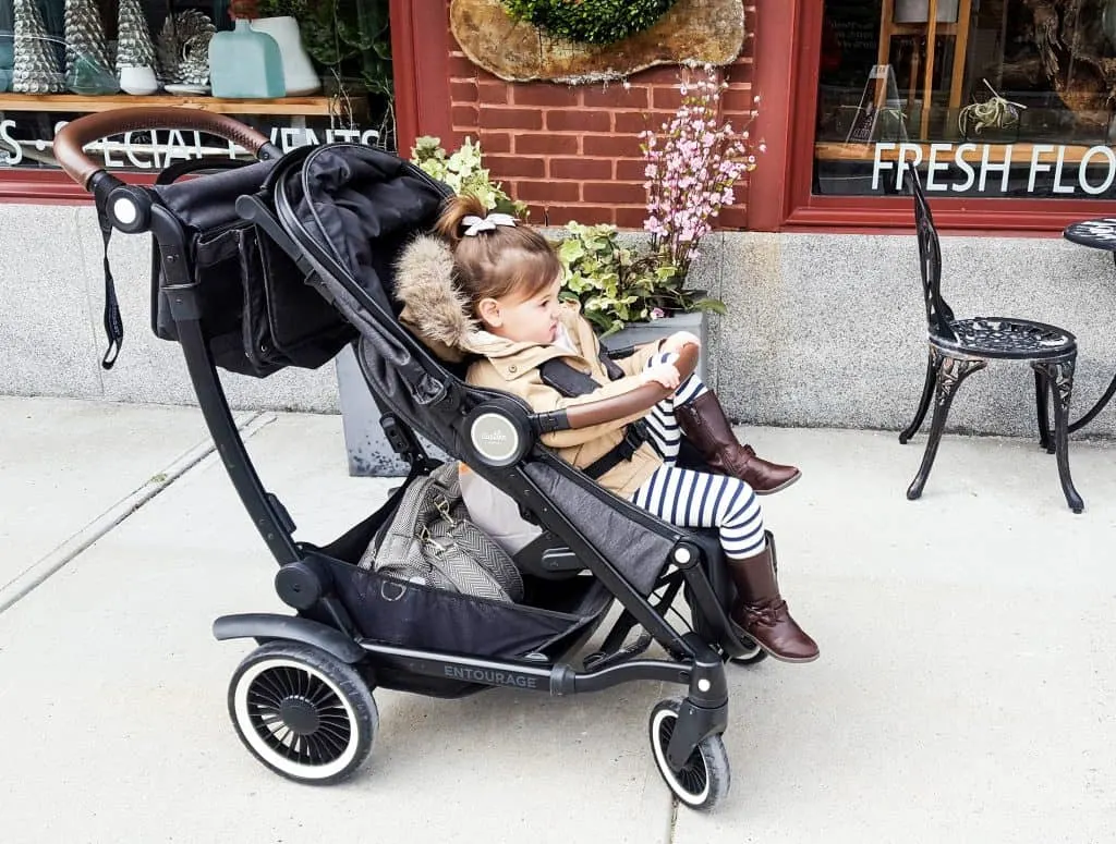 Toddler girl sits in travel stroller outside shops.