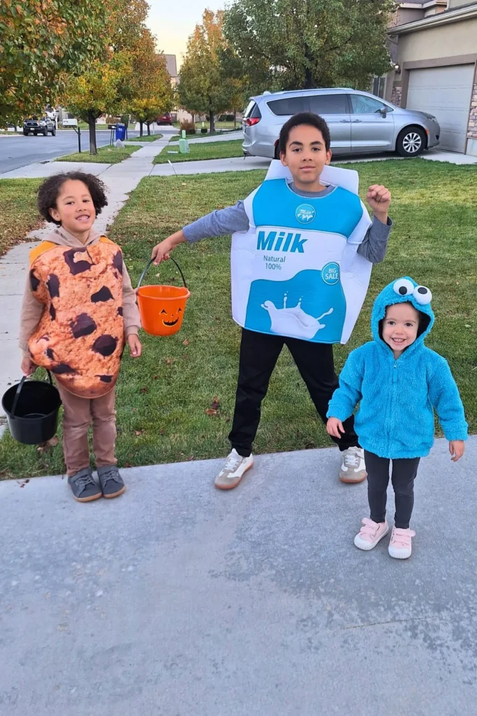 Three siblings dressed in a cookie costume, milk carton costume, and Cookie Monster costume for Halloween, standing on a driveway outside.