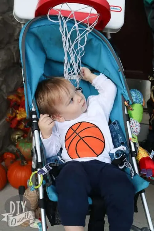 A toddler in a stroller with an image of a basketball on his shirt and a toy basketball hoop overhead.