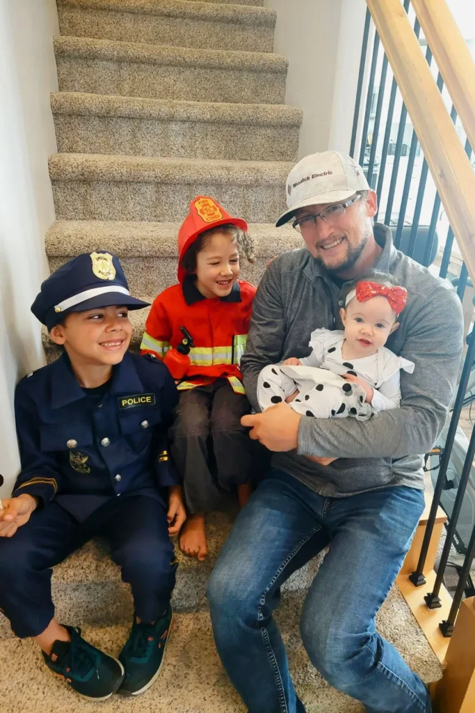 Children dressed as a police man firefighter, and a Dalmatian, sitting on the stairs with their dad.