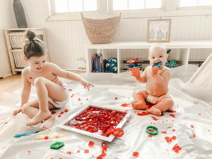 Young girl and baby play with red Jello over drop cloth.