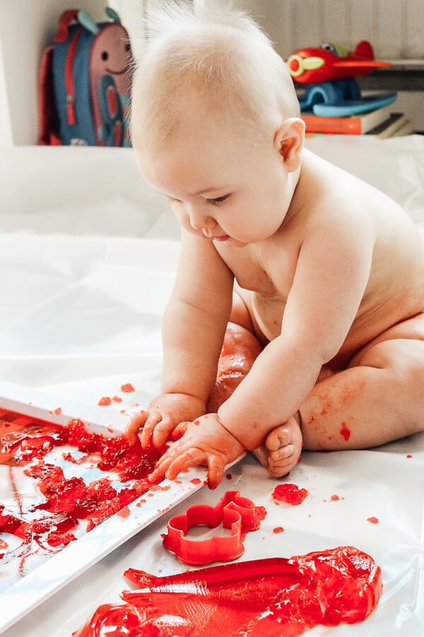 Baby uses hands to mash red Jello during sensory play.