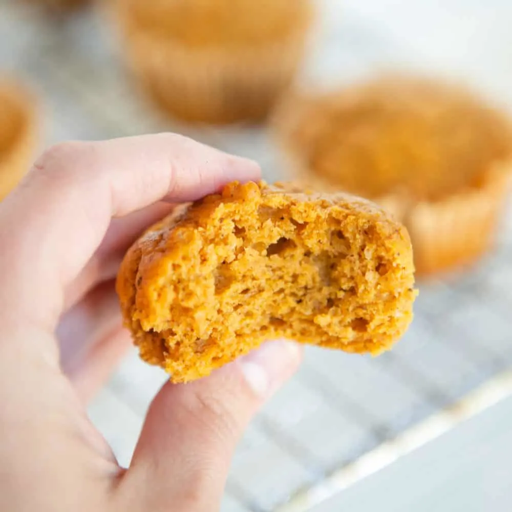A woman holds up a sweet potato muffin from a cooling rack with a bite taken out of it.