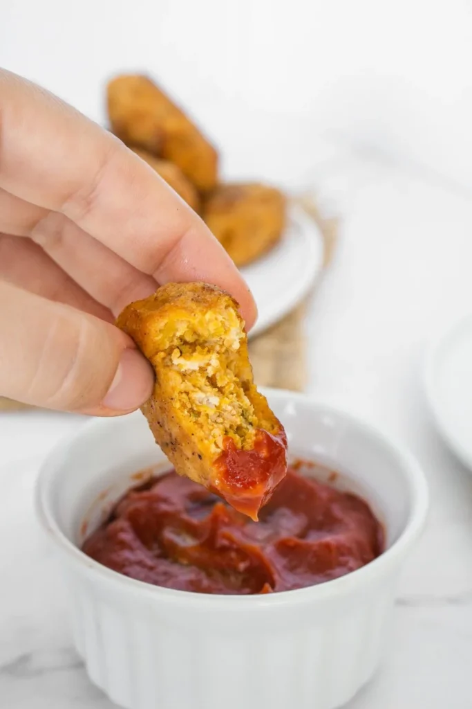 A woman lifts a sweet potato chicken nugget out of a dipping container with ketchup in it.