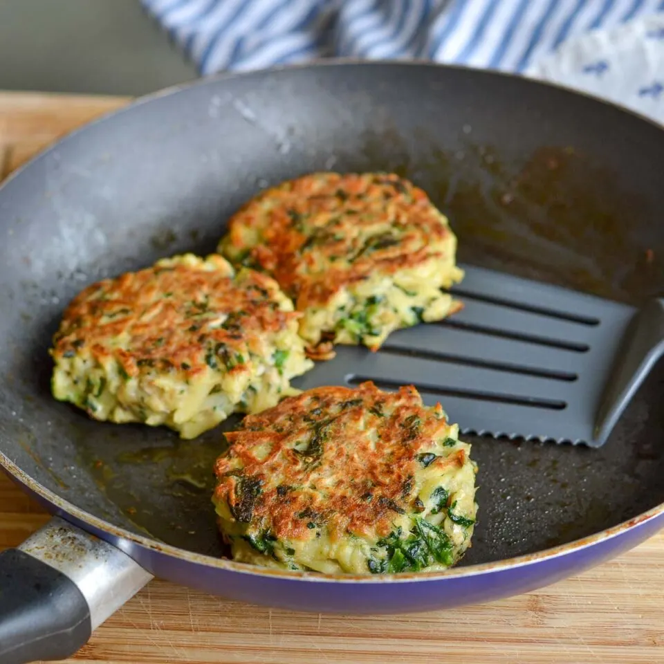 Hash brown and spinach salmon patties cooking in a skillet with a spatula.