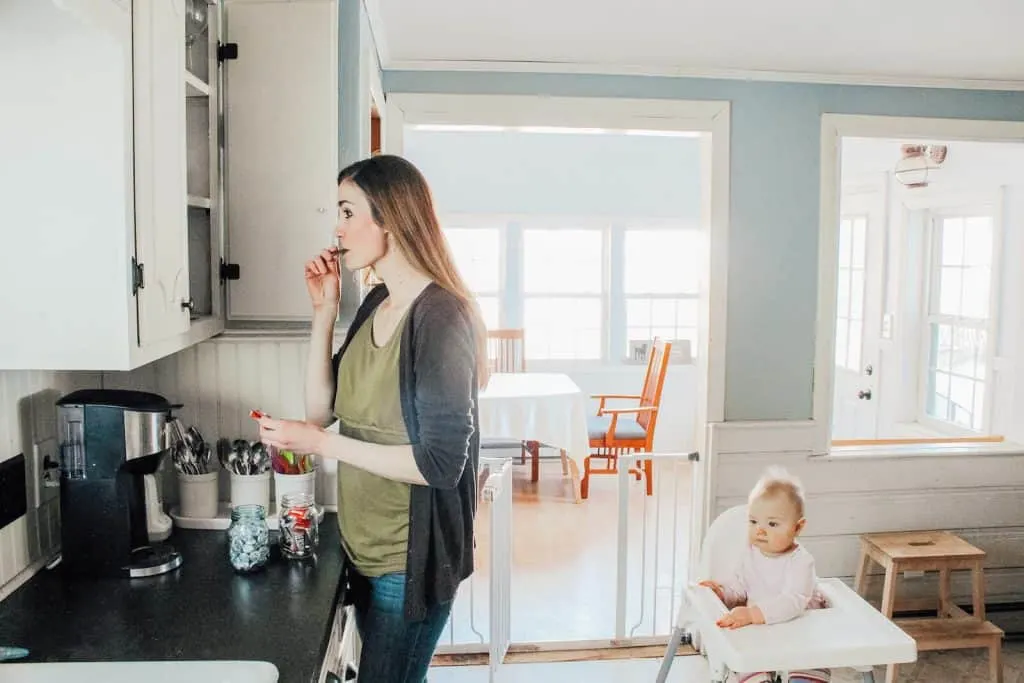 Mom eats chocolate in kitchen next to baby in high chair.