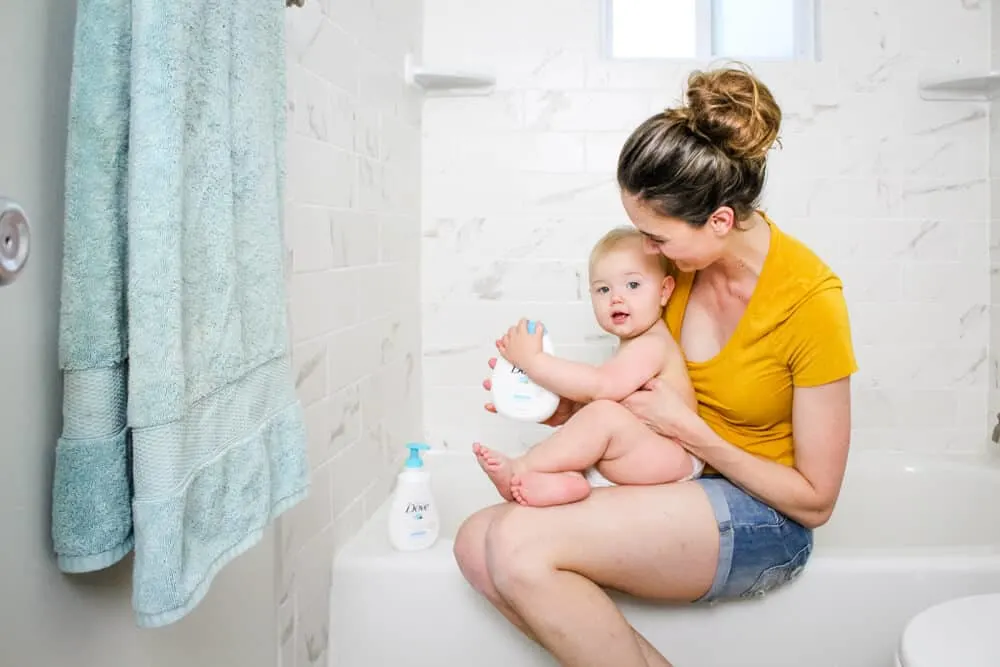 Mother holds baby on ledge of tub with baby skincare items.