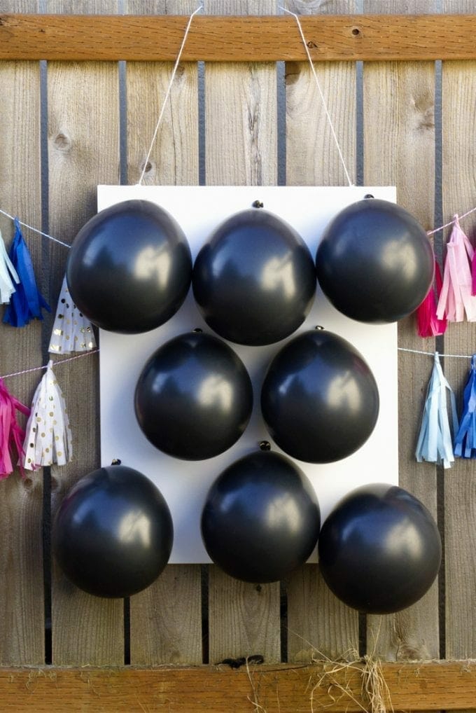 Black balloons filled with paint on a white canvas, hanging on a fence with pink and blue streamer garland.