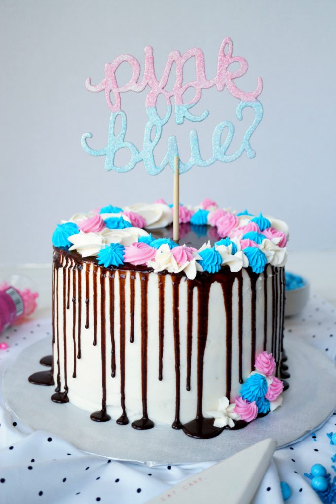 A white cake with chocolate drizzle and pink and blue decorations with a "Pink or Blue" cake topper.