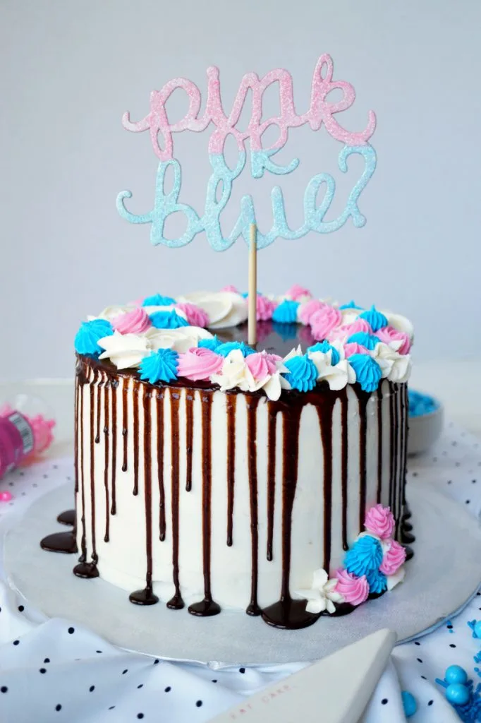 A white cake with chocolate drizzle and pink and blue decorations with a "Pink or Blue" cake topper.