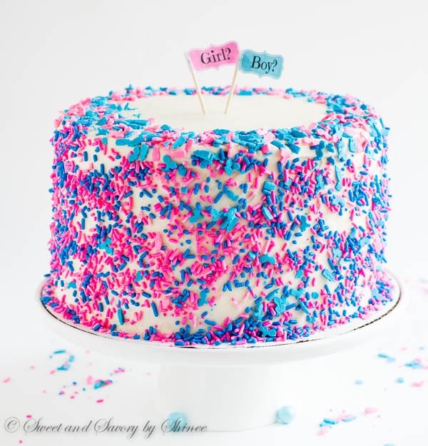 A white cake on a cake stand with pink and blue sprinkles a tiny signs that say boy and girl.