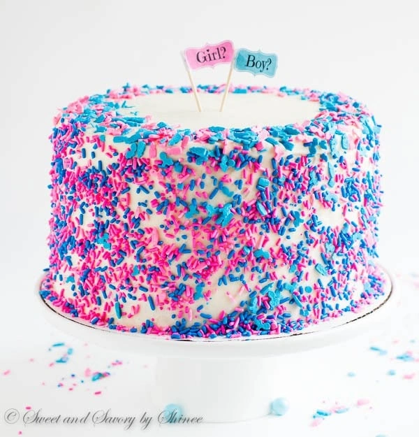A white cake on a cake stand with pink and blue sprinkles a tiny signs that say boy and girl.