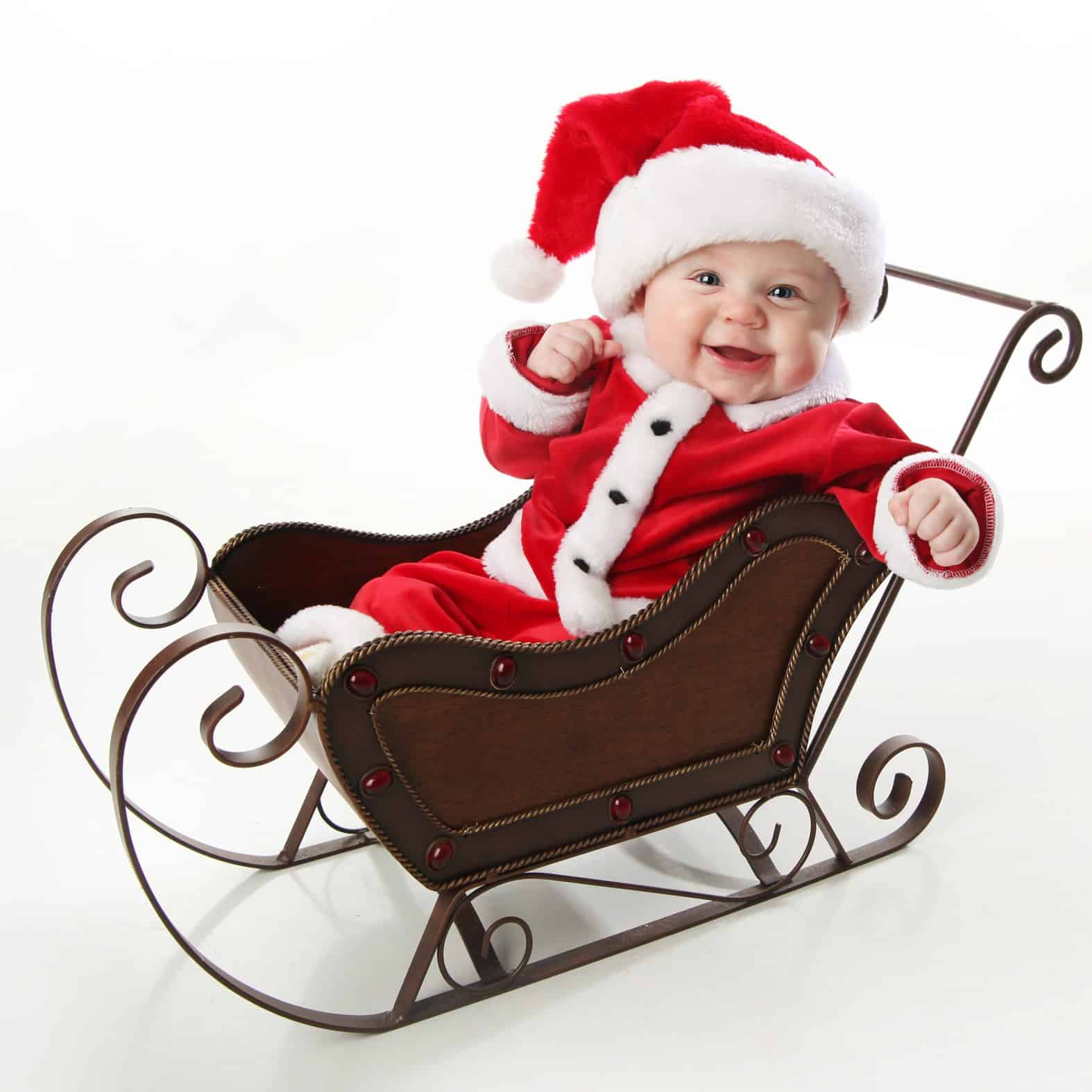 Adorable young baby wearing a santa claus suit and hat sitting in a metal Christmas snow sleigh