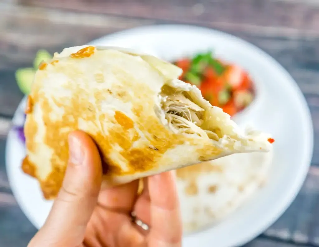 A women holds a chicken quesadilla with out bite taken out of it over a dinner plate.