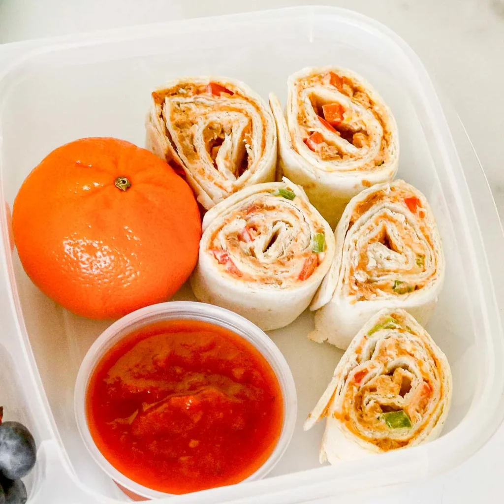 Taco pinwheels in a lunch container with an orange and dipping sauce.