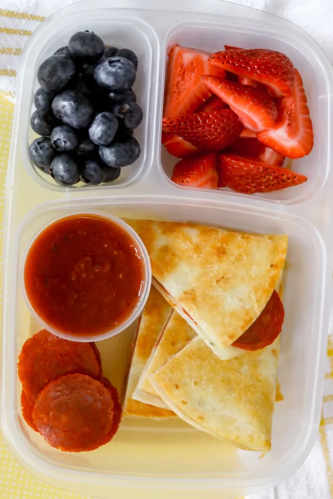 Pizza quesadillas in a lunch container with dipping sauce and fruit.