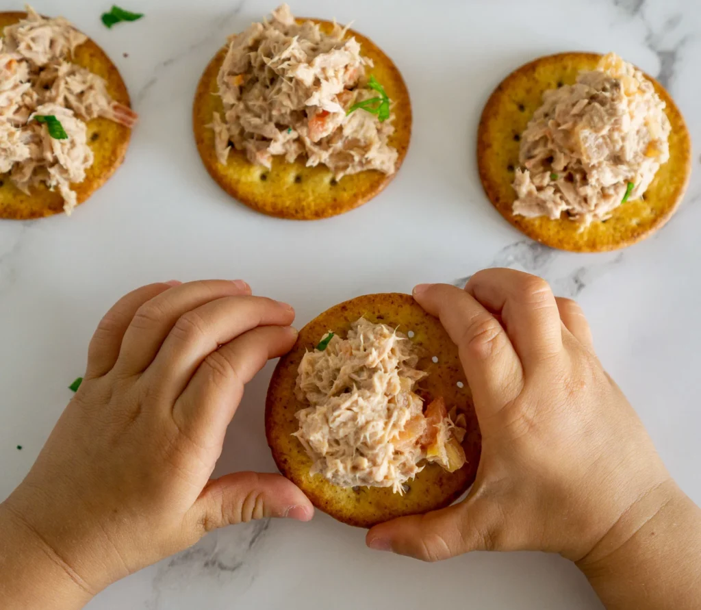 A toddler picks up a cracker with tuna salad on it.