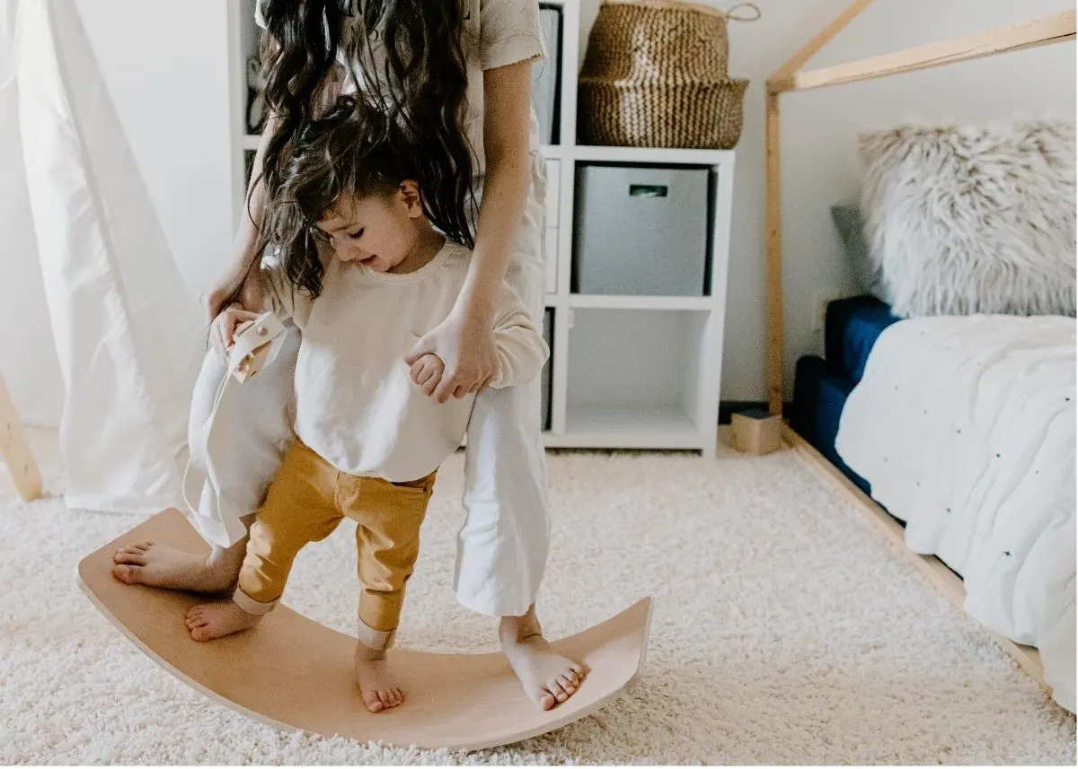 A mother helps her toddler play on a wooden Montessori rocking board.