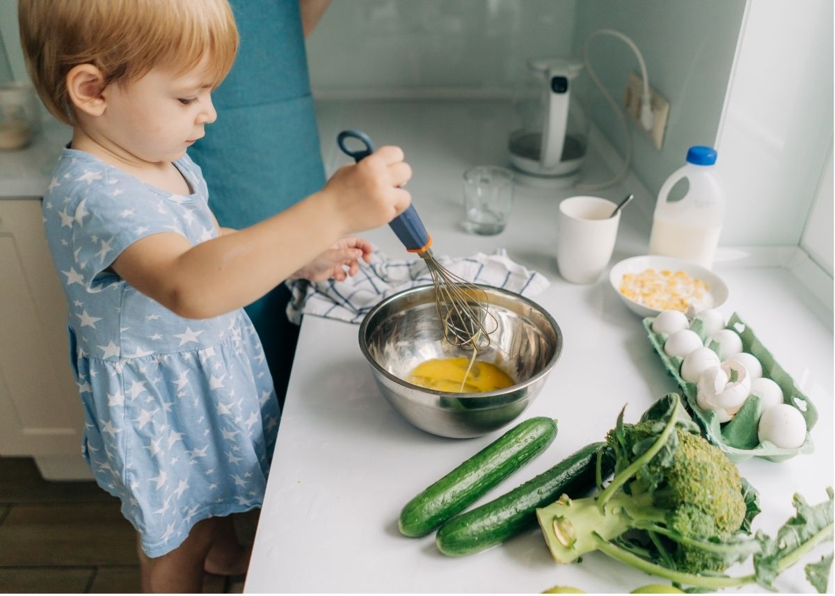 A toddler stirs eggs in a metal bowl with a whisk.