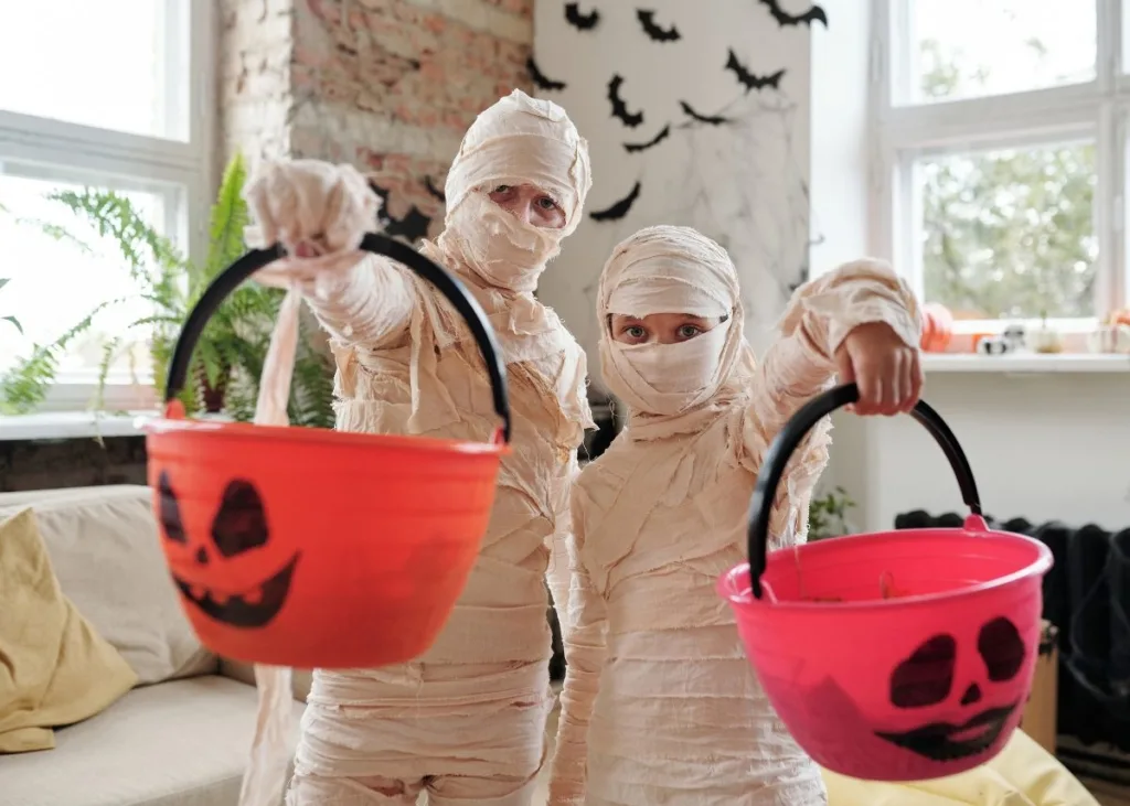 Two people dressed up as mummies holding pumpkin buckets with candy inside.