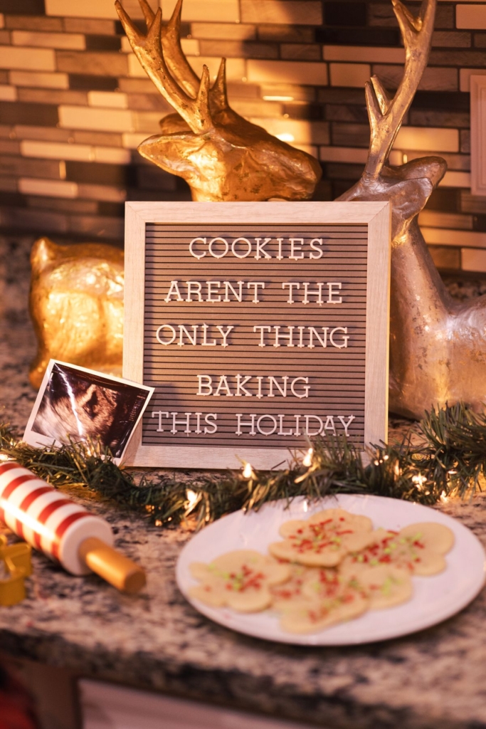 A pregnancy announcement sign on a kitchen counter with Christmas decorations and cookies.