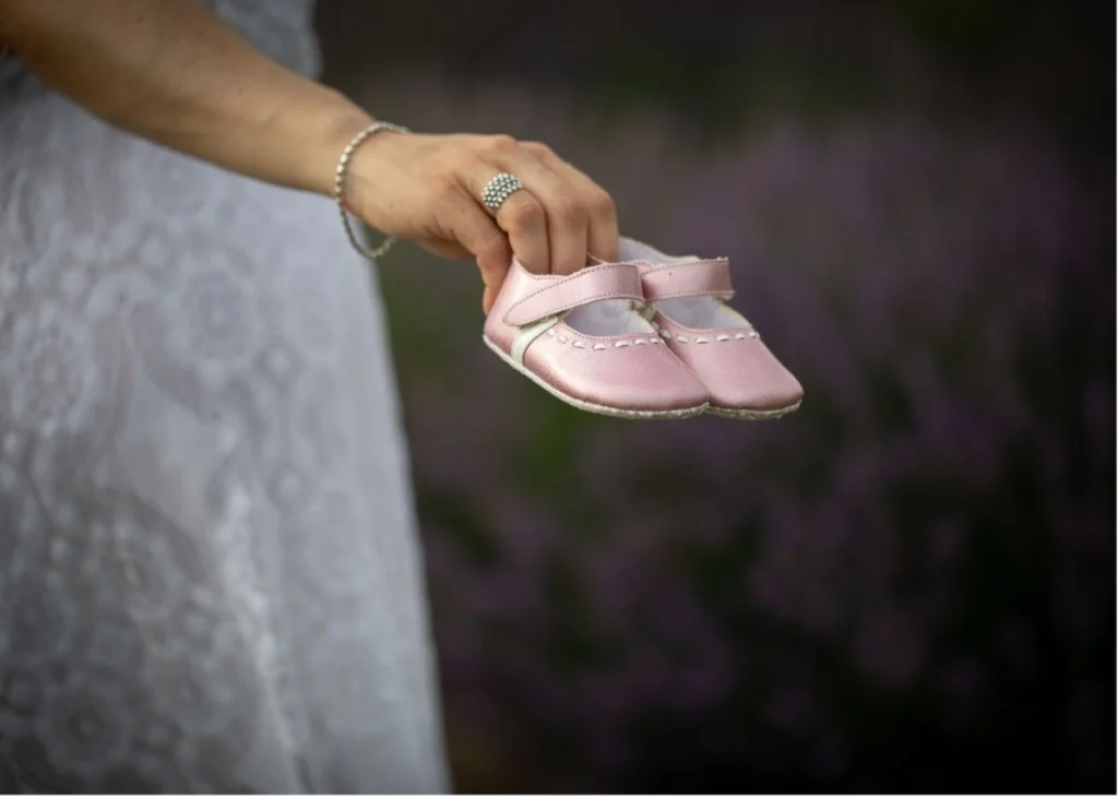 A woman in a white dress holds out pink baby shoes outside.