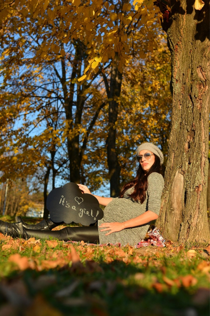 A woman holds a sign that says "it's a girl" while sitting against the trunk of a tree during fall.