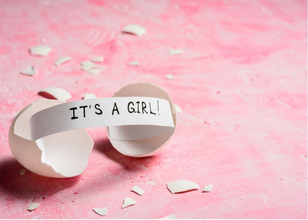 A note that says "It's a girl" popping out of a broken eggshell on a pink background.