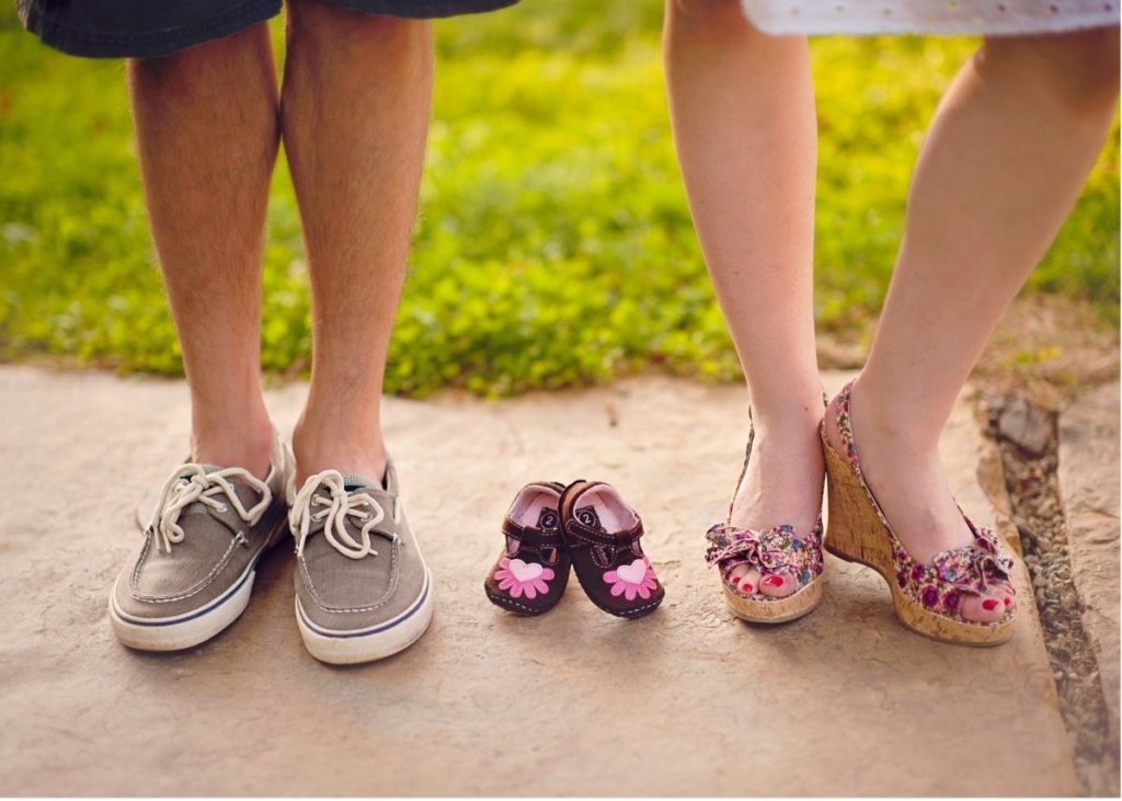 A couple stands outside with girl baby shoes in between them on the ground.