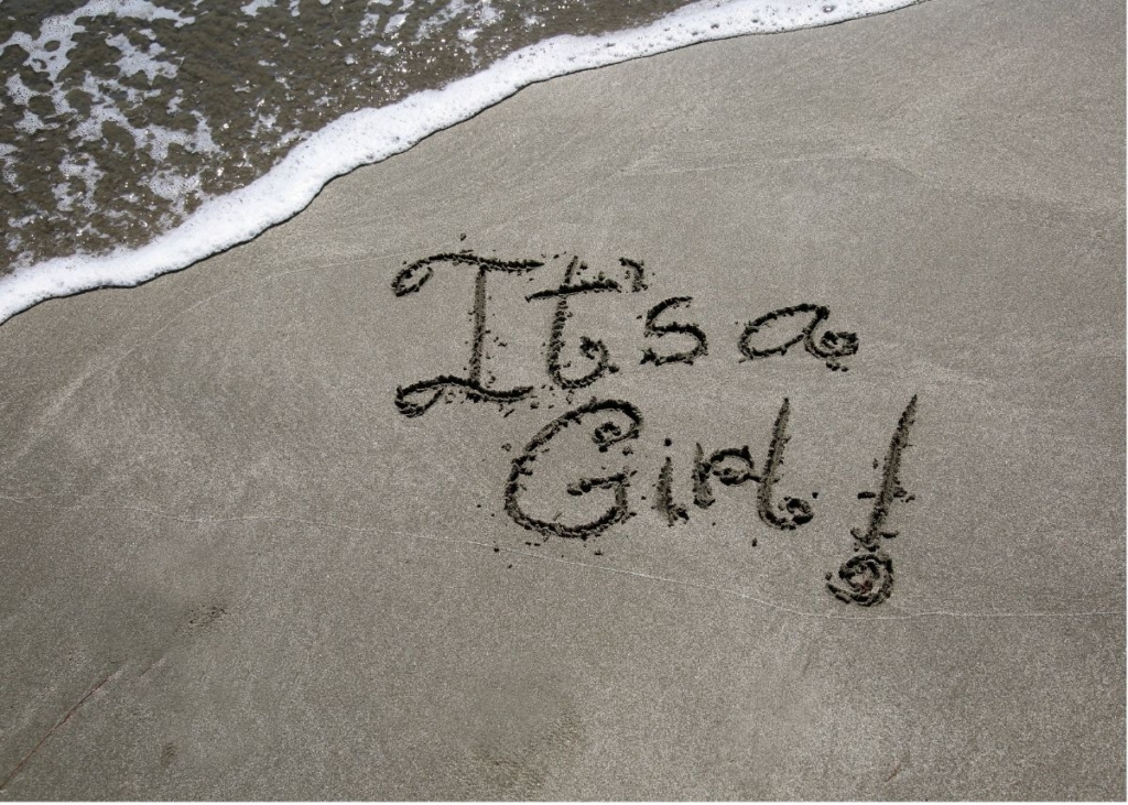 "It's a girl" written in the sand by the ocean.