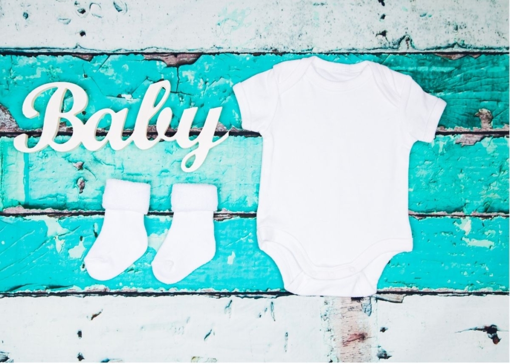 A white onesie and socks on blue distressed wood planks with a sign that says "baby".