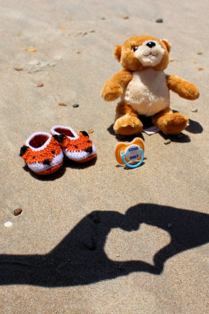 A couple makes a heart shadow with their hands at the beach in front of a teddy bear, pacifier, and baby shoes.