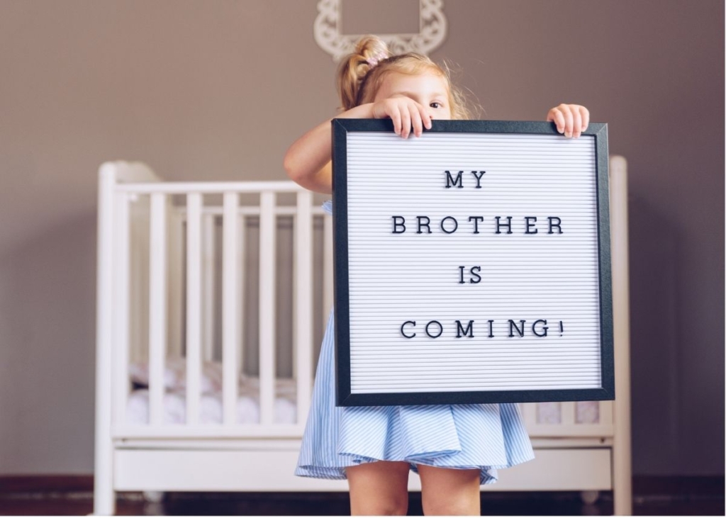 A little girl holds up a sign that says "My Brother is Coming" in a nursery.