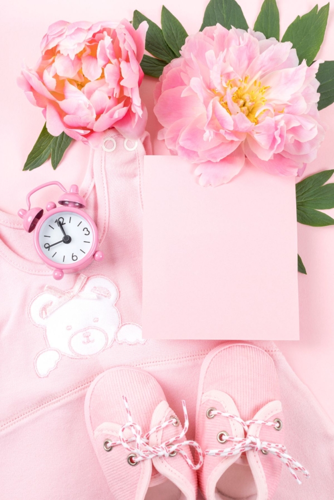Pink baby shoes next to a pink clock and spring flowers.