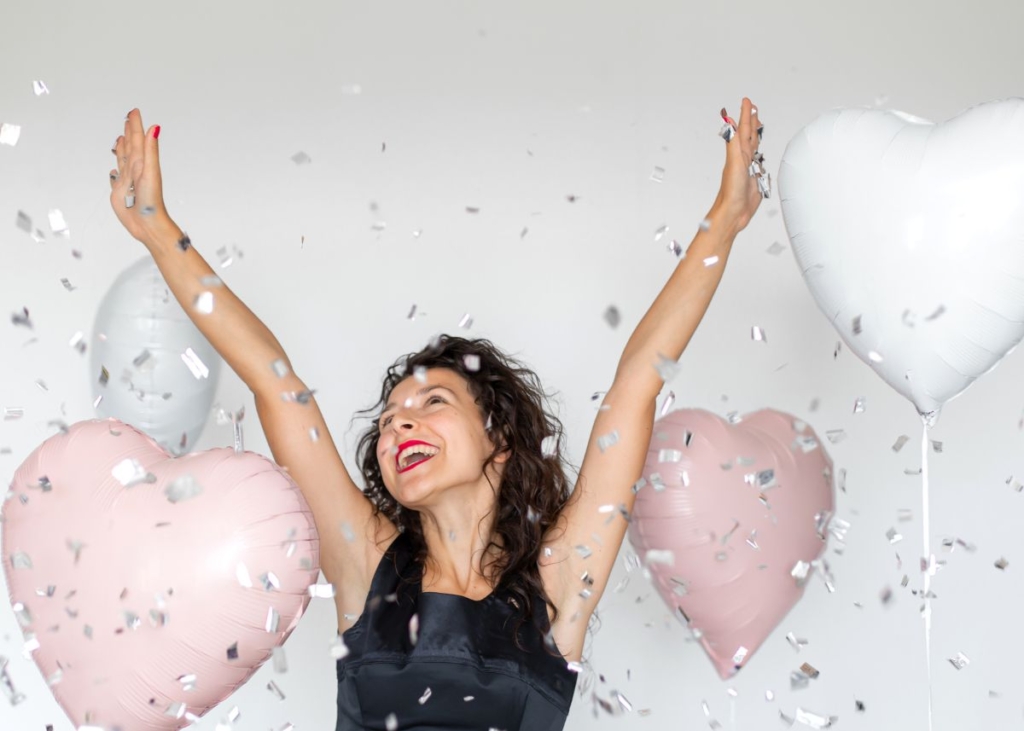 An excited woman throws her hands up with heart balloons and confetti in the background.
