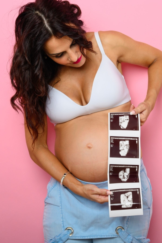 A pregnant woman holds up ultrasound photos in front of a pink background.