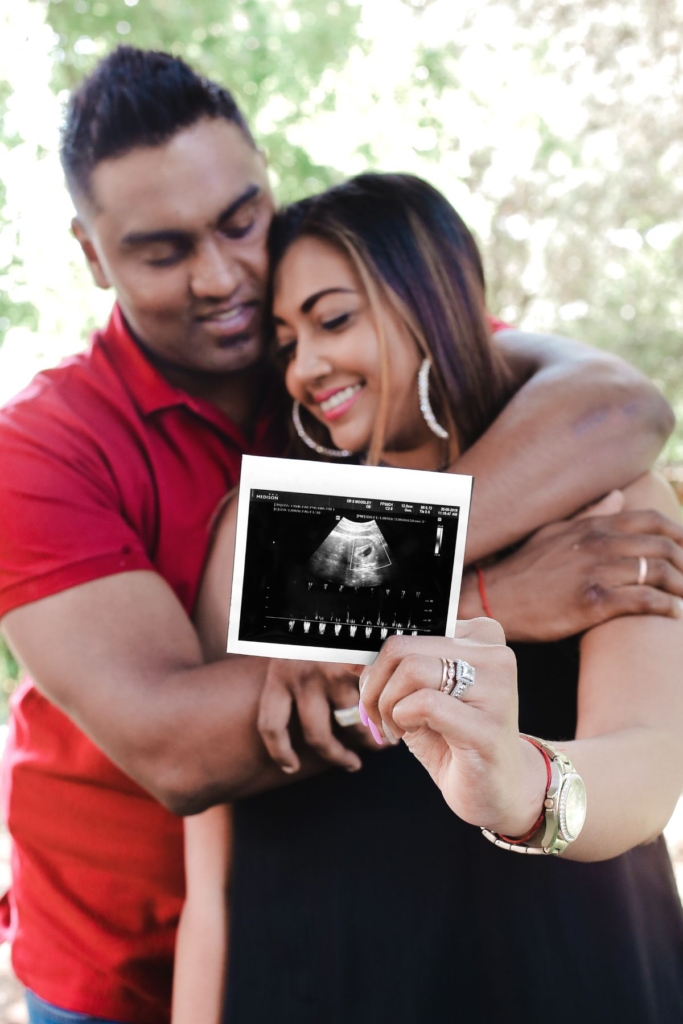 A couple embraces while holding up ultrasound photos outside.
