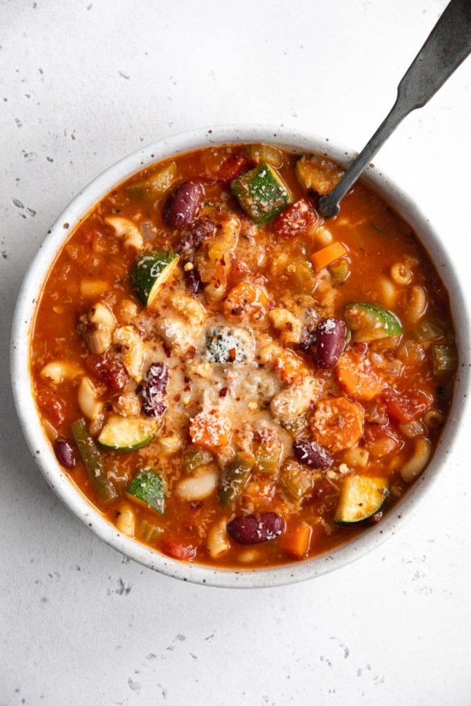 Minestrone in a white bowl with zucchini, beans, noodles, and other vegetables.