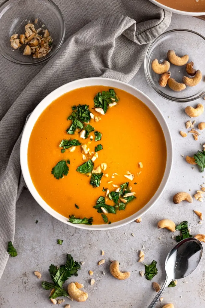 Carrot ginger soup in a white bowl with cashews and green garnish on top.