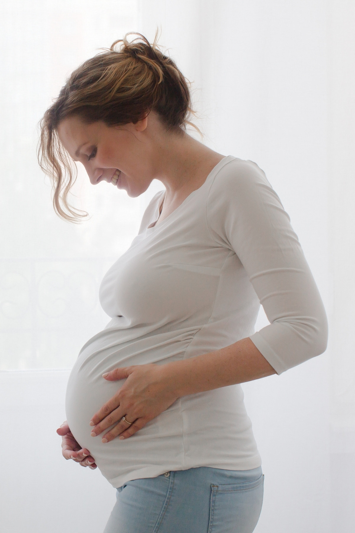 Pregnant woman cradles baby bump in front of a window shining with soft white light.