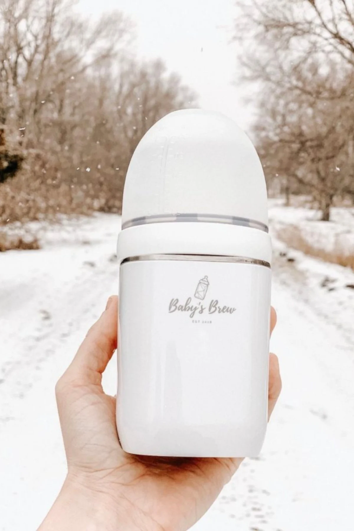 Woman holds up Baby's Brew baby bottle warmer in front of snowy, tree-lined road.