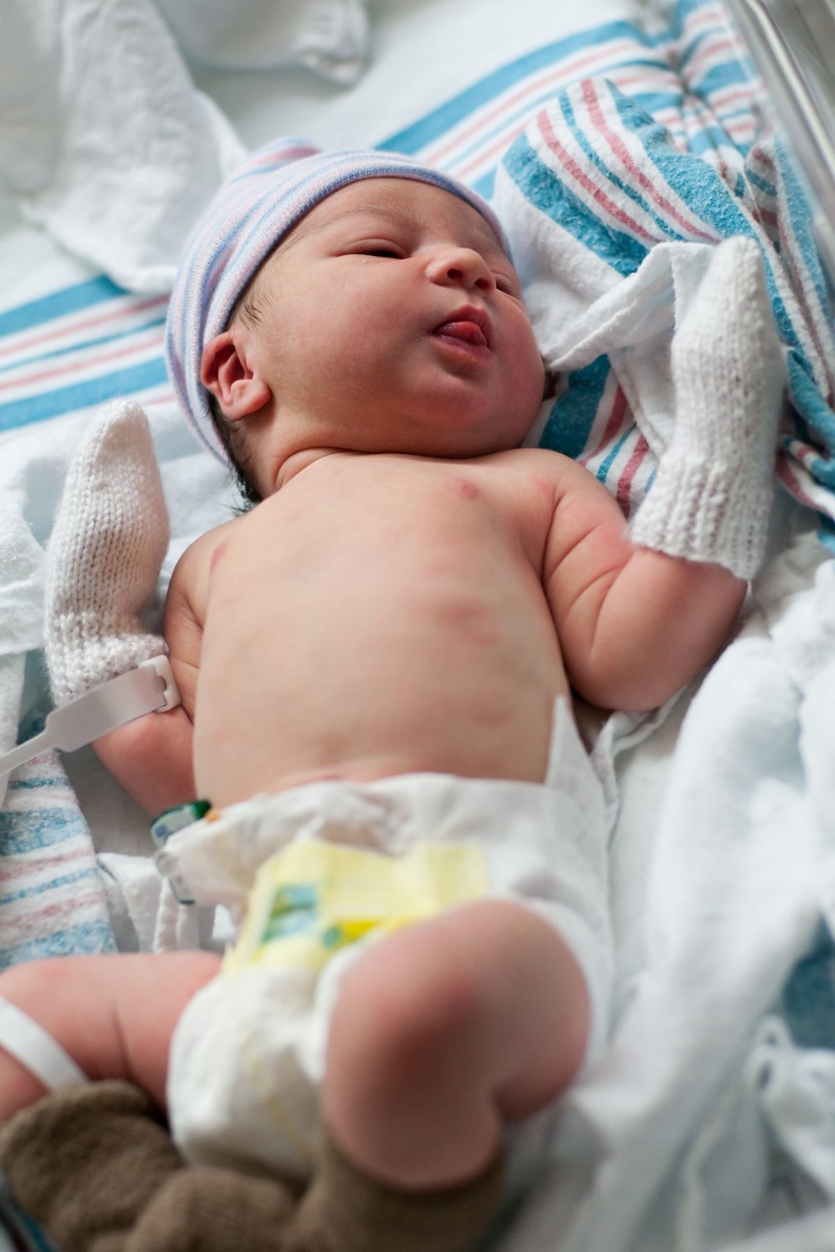 Newborn baby lays on striped blanket in hospital bassinet with mittens on hands.