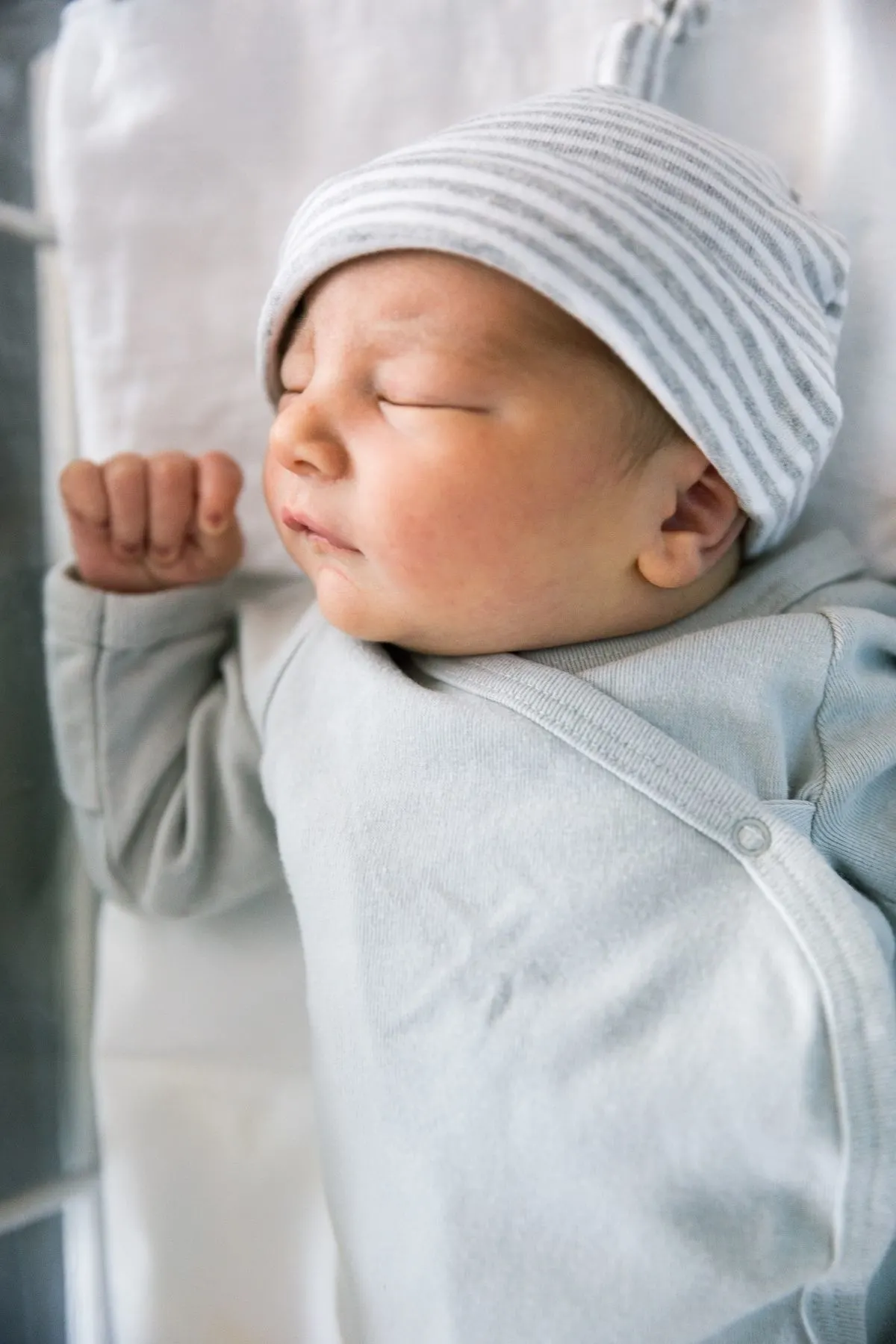 Newborn baby sleeps in hospital bed wearing a gray and white onesie and striped hat.