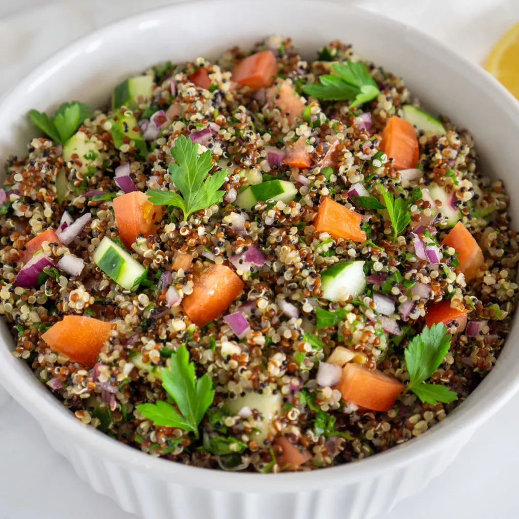 A grain salad with cucumbers, tomatoes, and green garnish in a white bowl.