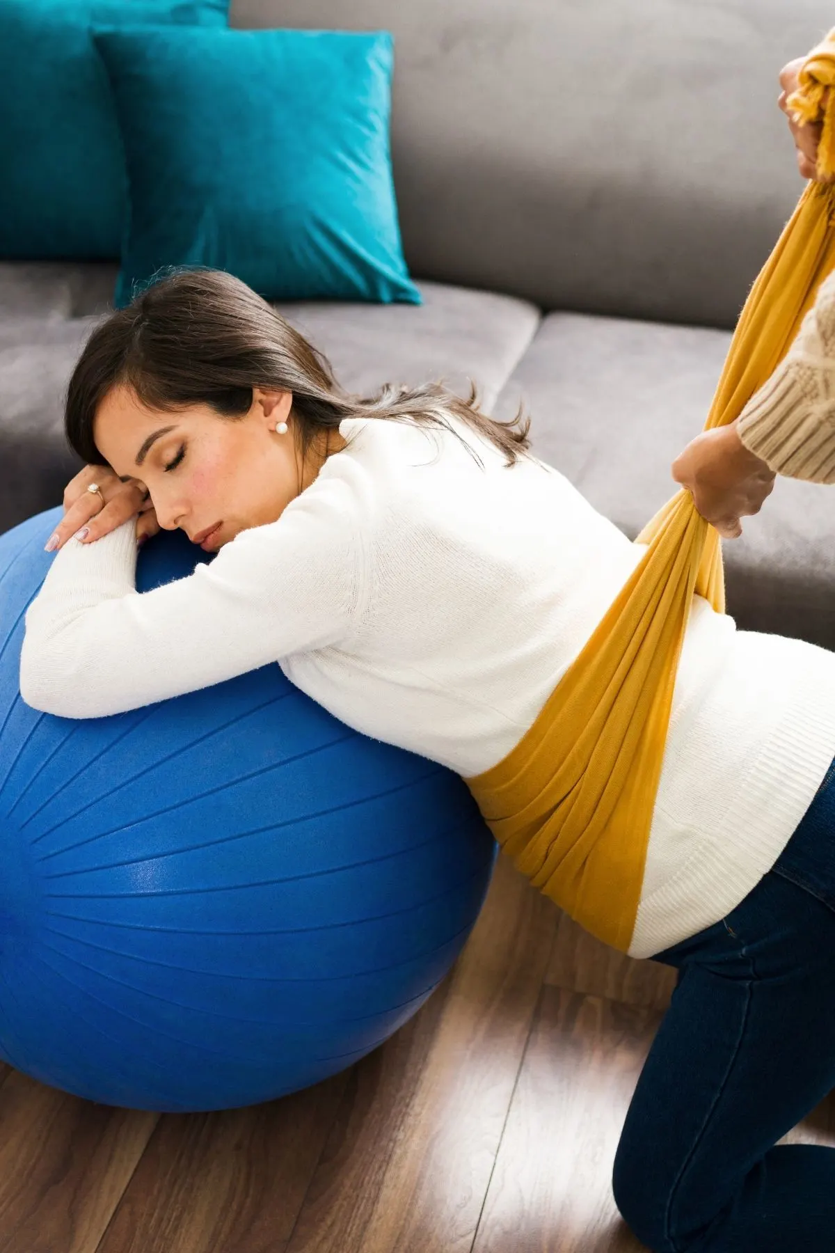 Woman leans over birthing ball while her midwife relieves her stomach pain with rebozo scarf.