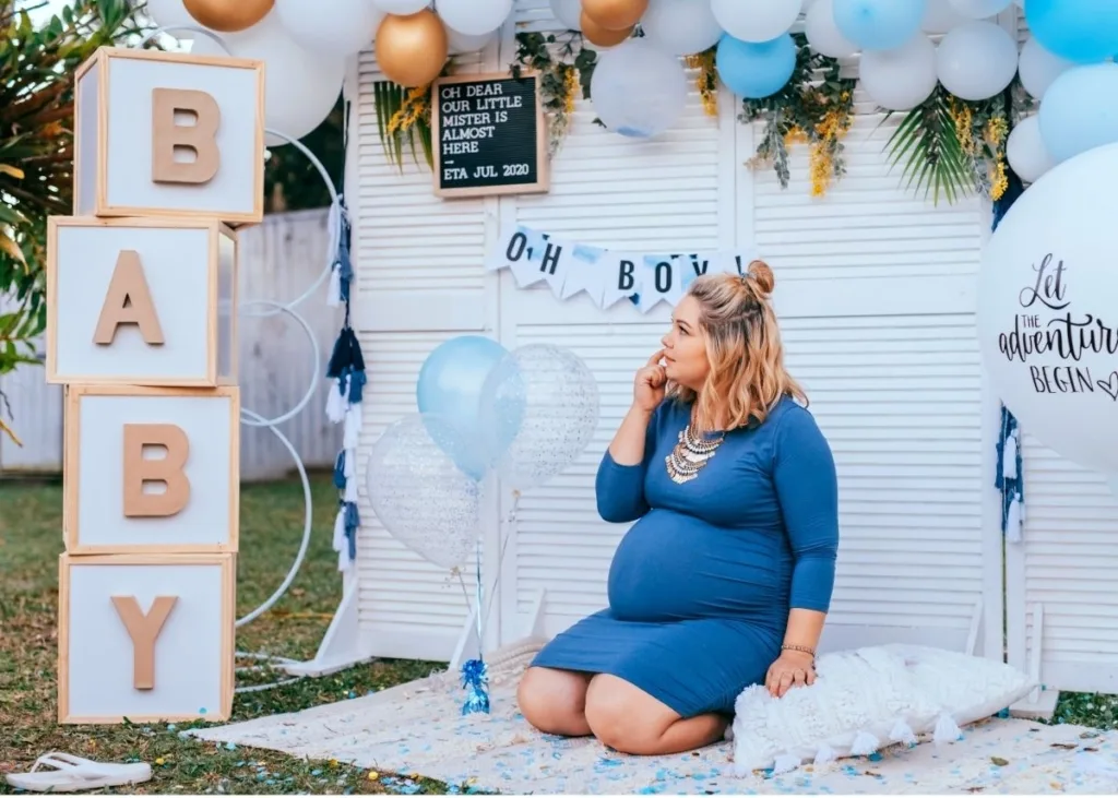 A woman sits in front of a white backdrop outside, decorated for a boy baby shower.