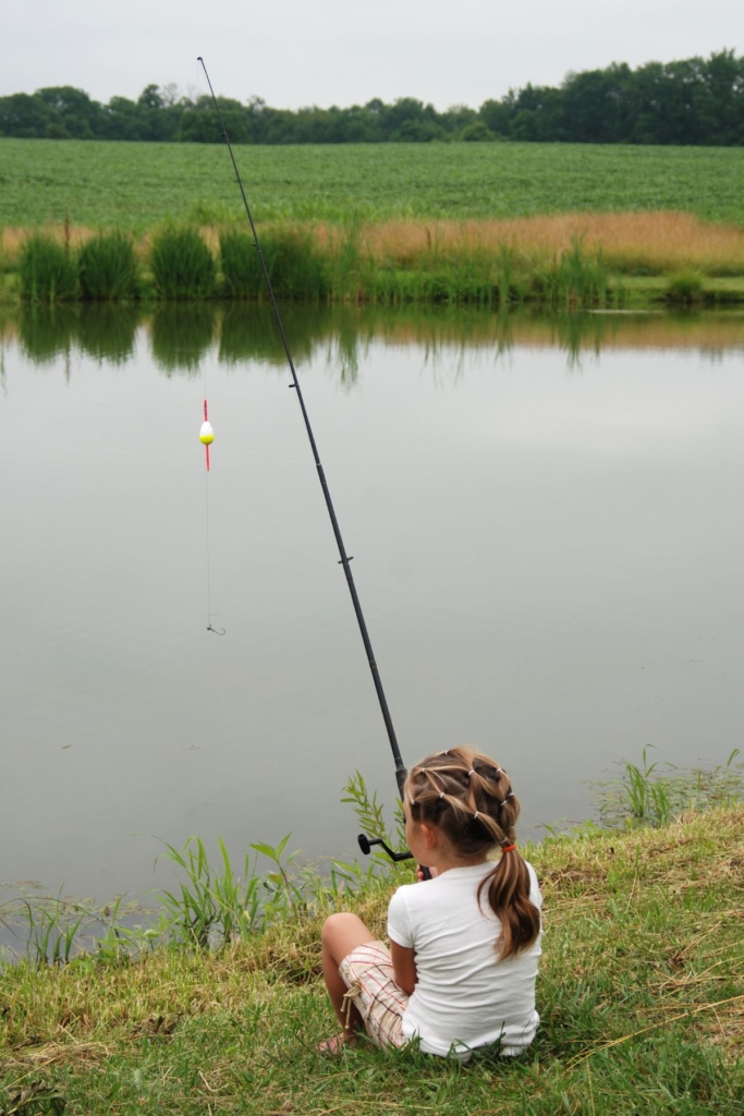 A little girl sits on the bank of a pond with a fishing rod in the water.