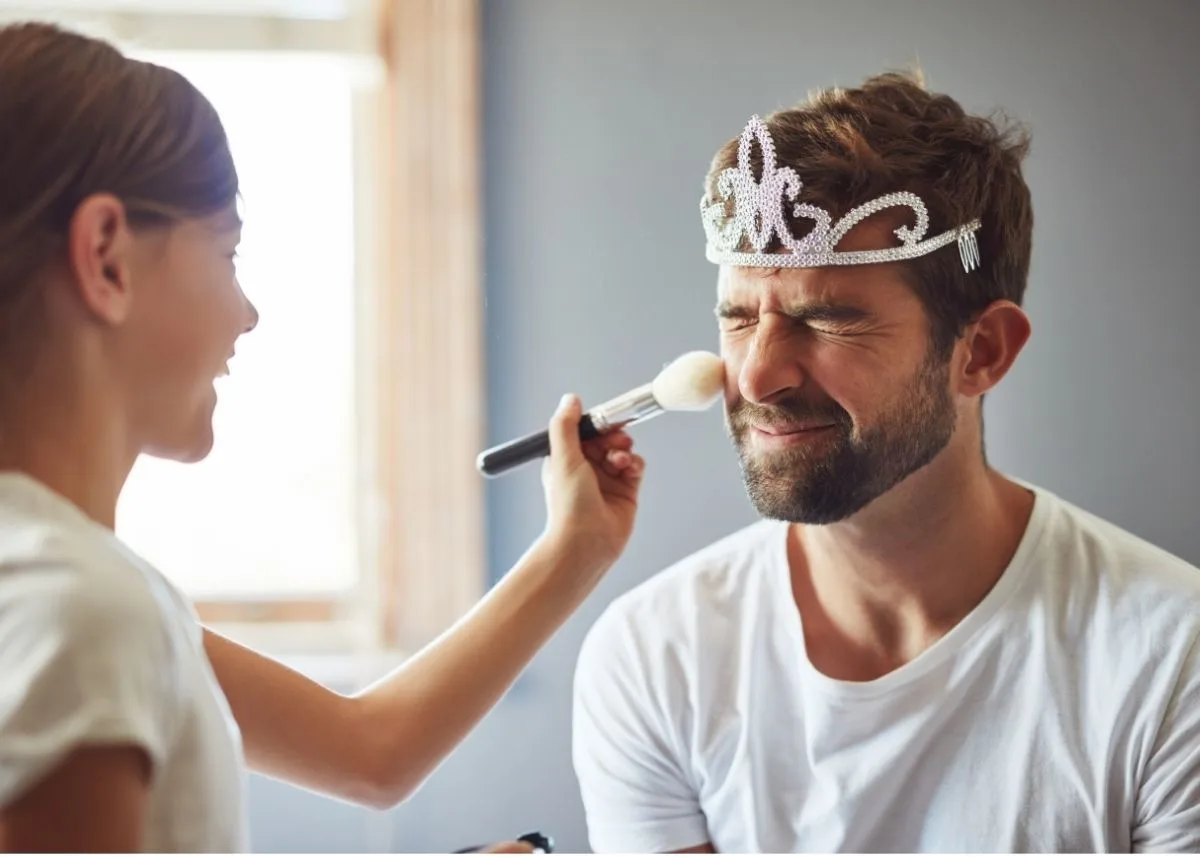 A young girl applies makeup to her dad's face who's wearing a crown.